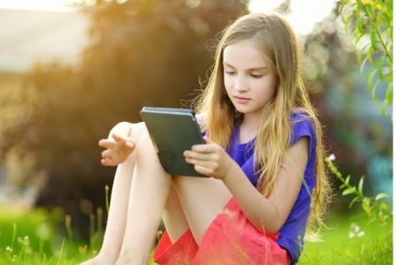 Meisje zit op grasveld. Zij leest een boek met behulp van een tablet.