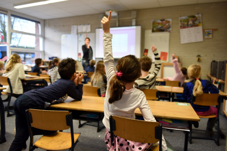 Een volle klas met kinderen. Één kind steekt de vinger hoog in de lucht op.