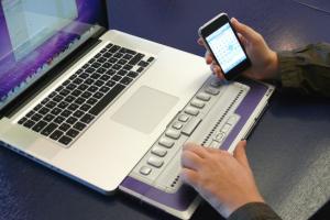 Een man bedient zijn laptop met brailleleesregel en heeft een smartphone in de andere hand