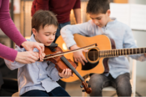 Een leerling krijgt les en speelt op een viool. Ernaast zit nog een leerling, deze speelt op een gitaar.
